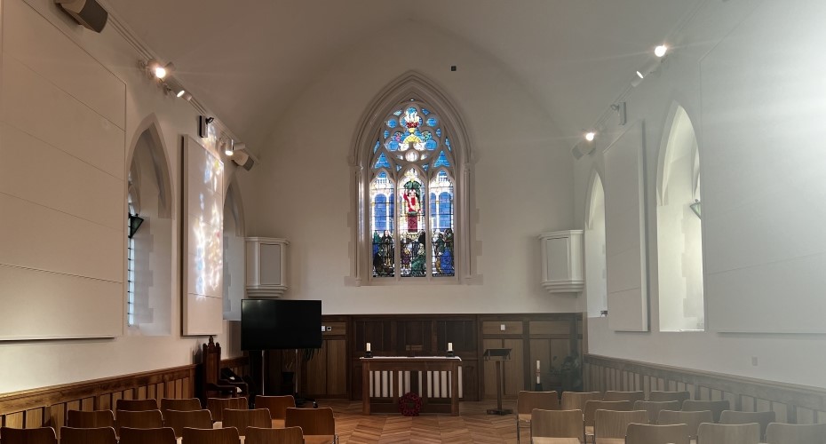 The interior of St Luke's Chapel, facing the altar. Showing white walls, a parquet wooden floor and a predominantly blue stained-glass window.